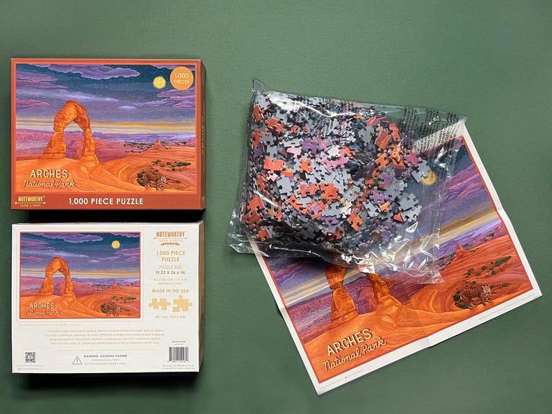 May include: A 1,000 piece jigsaw puzzle featuring a scenic view of Delicate Arch in Arches National Park, Utah. The puzzle box shows a colorful sunset sky with a red rock archway. The puzzle is made in the USA.
