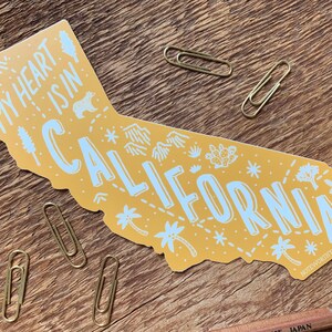 May include: Yellow sticker shaped like the state of California with the text "My heart is in California" and various illustrations of mountains, palm trees, cacti, and stars.