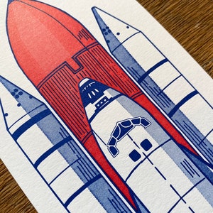 May include: A white and blue illustration of a space shuttle with a red rocket booster attached. The shuttle has a detailed design with multiple windows and a hatch.