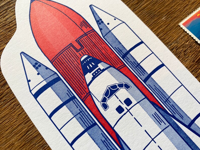 May include: A white and blue illustration of a space shuttle with a red rocket booster attached. The shuttle has a detailed design with multiple windows and a hatch.