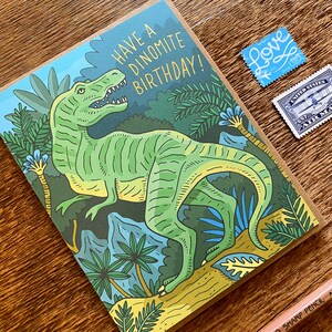 Op de afbeelding: Een groene dinosaurus met gele strepen staat in een jungle-omgeving. De kaart luidt "Have a Dinomite Birthday!"