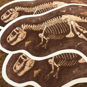 Peut inclure: Trois autocollants blancs sur fond brun repr&eacute;sentant une illustration de squelette de dinosaure. Le dinosaure est repr&eacute;sent&eacute; de profil, la t&ecirc;te tourn&eacute;e vers la gauche. Le squelette est d&eacute;taill&eacute; et complexe, avec des c&ocirc;tes, des vert&egrave;bres et des membres visibles. L'arri&egrave;re-plan est brun fonc&eacute;, ressemblant &agrave; de la terre ou de la roche, avec une texture subtile.