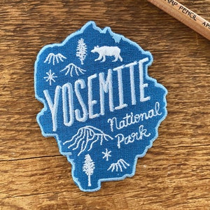 Könnte beinhalten: Ein blauer gestickter Aufnäher mit dem Text "Yosemite National Park" in Weiß. Der Aufnäher zeigt eine stilisierte Bergkette, einen Bären und eine Kiefer.