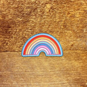 Pu&ograve; includere: Una toppa ricamata a forma di arcobaleno con un contorno blu. L'arcobaleno ha sette colori: rosso, arancione, giallo, verde, blu, indaco e violetto.