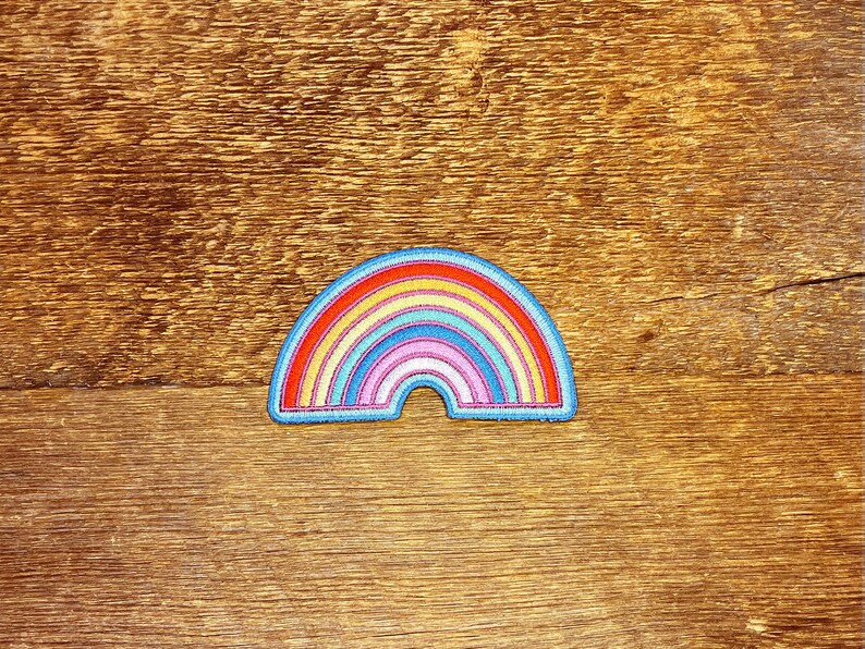 Pu&ograve; includere: Una toppa ricamata a forma di arcobaleno con un contorno blu. L'arcobaleno ha sette colori: rosso, arancione, giallo, verde, blu, indaco e violetto.