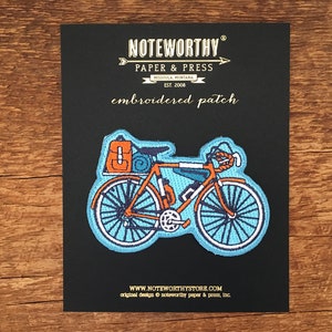 May include: Embroidered patch of a blue bicycle with orange luggage strapped to the back. The patch is on a black background with the text "NOTEWORTHY PAPER & PRESS MISSOULA MONTANA EST. 2008 embroidered patch WWW.NOTEWORTHYSTORE.COM original design noteworthy paper & press, Inc."