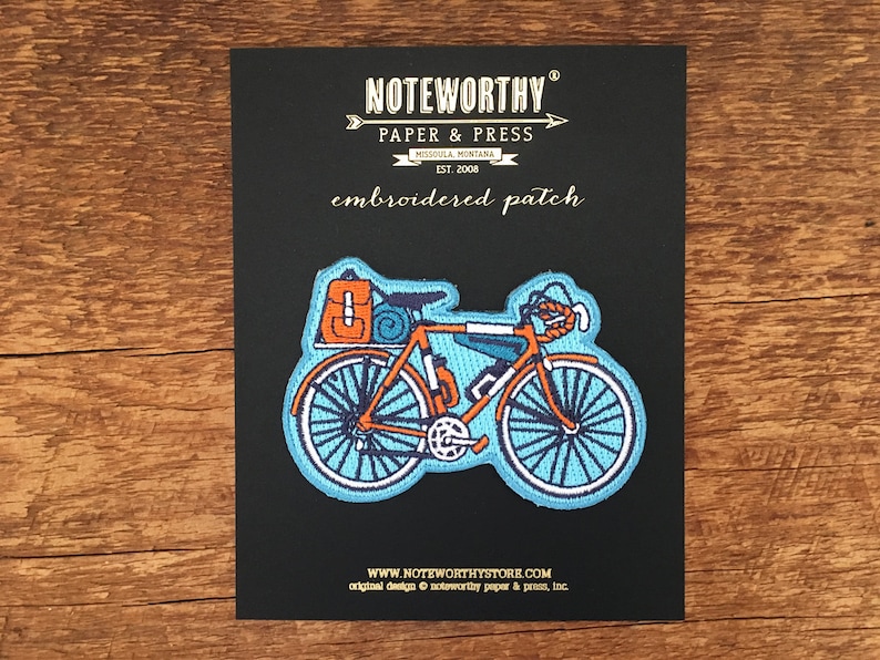 May include: Embroidered patch of a blue bicycle with orange luggage strapped to the back. The patch is on a black background with the text "NOTEWORTHY PAPER & PRESS MISSOULA MONTANA EST. 2008 embroidered patch WWW.NOTEWORTHYSTORE.COM original design noteworthy paper & press, Inc."
