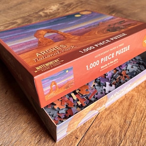 May include: A 1,000 piece jigsaw puzzle of Arches National Park in Utah. The box features a colorful image of Delicate Arch, a natural sandstone arch formation. The puzzle pieces are scattered inside the box.