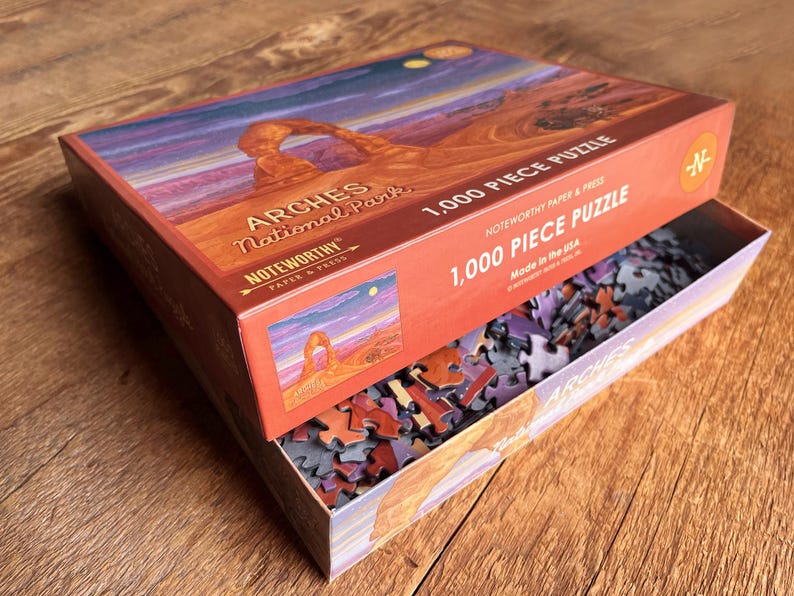 May include: A 1,000 piece jigsaw puzzle of Arches National Park in Utah. The box features a colorful image of Delicate Arch, a natural sandstone arch formation. The puzzle pieces are scattered inside the box.