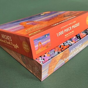 May include: A 1,000 piece jigsaw puzzle of Arches National Park in Utah. The box features a colorful illustration of Delicate Arch, a natural sandstone arch formation. The puzzle is made in the USA.
