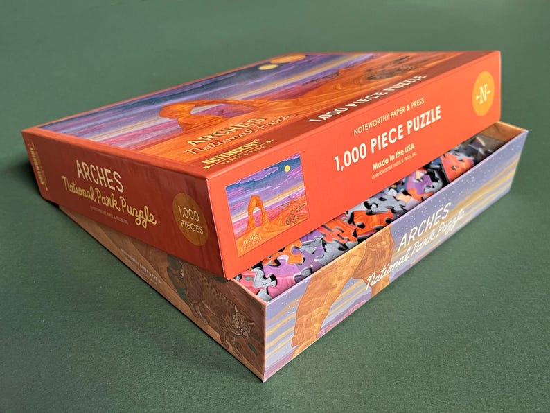 May include: A 1,000 piece jigsaw puzzle of Arches National Park in Utah. The box features a colorful illustration of Delicate Arch, a natural sandstone arch formation. The puzzle is made in the USA.