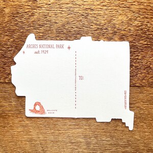 May include: A white postcard with a red outline of Delicate Arch in Arches National Park, Utah. The postcard says "Arches National Park est. 1929" and "Delicate Arch" in red text. The back of the postcard has a space for a message, an address, and a stamp.