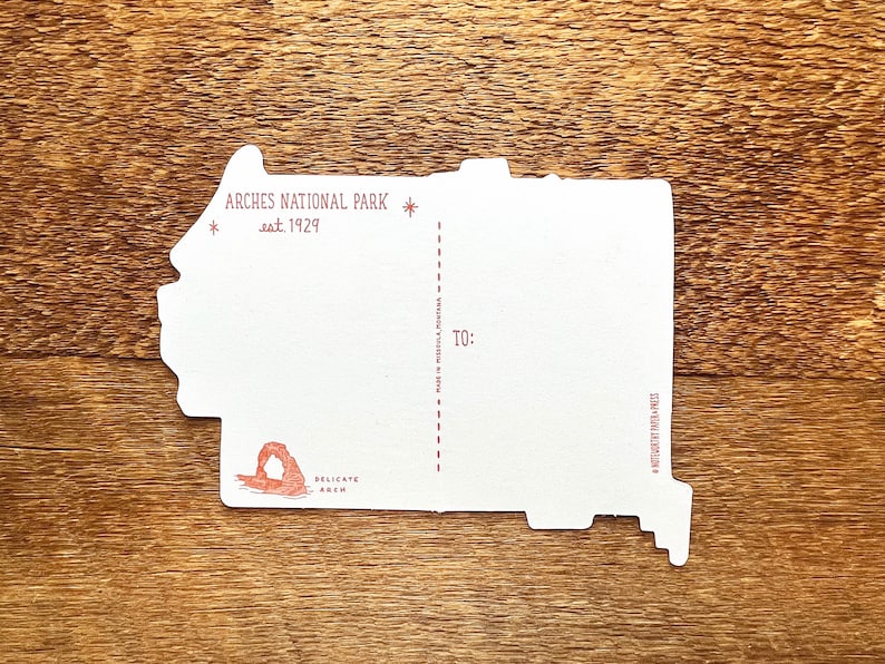 May include: A white postcard with a red outline of Delicate Arch in Arches National Park, Utah. The postcard says "Arches National Park est. 1929" and "Delicate Arch" in red text. The back of the postcard has a space for a message, an address, and a stamp.