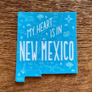 May include: A blue sticker with a white outline of the state of New Mexico. The sticker says "My heart is in New Mexico" with a map-like design inside the state outline. The design includes mountains, a sun, a cactus, and a roadrunner.