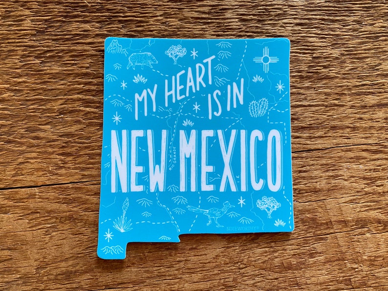 May include: A blue sticker with a white outline of the state of New Mexico. The sticker says "My heart is in New Mexico" with a map-like design inside the state outline. The design includes mountains, a sun, a cactus, and a roadrunner.