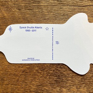 May include: White card shaped like a space shuttle with blue stars and the text "Space Shuttle Atlantis 1985-2011" printed on the top. The card has a dashed line down the middle and the text "TO:" and "MADE IN MISSOULA, MONTANA" printed on the right side. The card is printed by "NOTEWORTHY PAPER & PRESS".