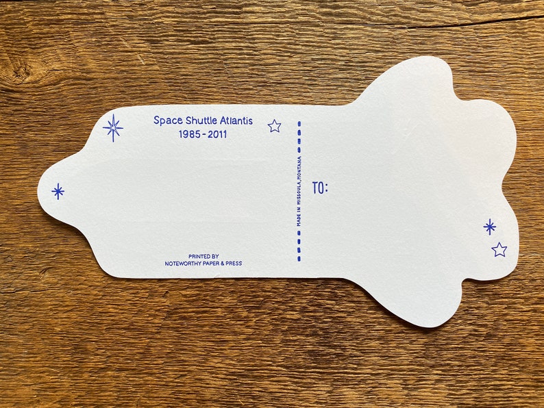 May include: White card shaped like a space shuttle with blue stars and the text "Space Shuttle Atlantis 1985-2011" printed on the top. The card has a dashed line down the middle and the text "TO:" and "MADE IN MISSOULA, MONTANA" printed on the right side. The card is printed by "NOTEWORTHY PAPER & PRESS".