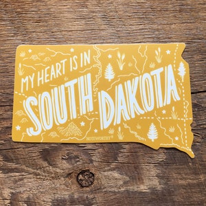 Pu&ograve; includere: Adesivo giallo a forma dello stato del Dakota del Sud con il testo "My heart is in South Dakota" e la parola "Noteworthy" in basso.