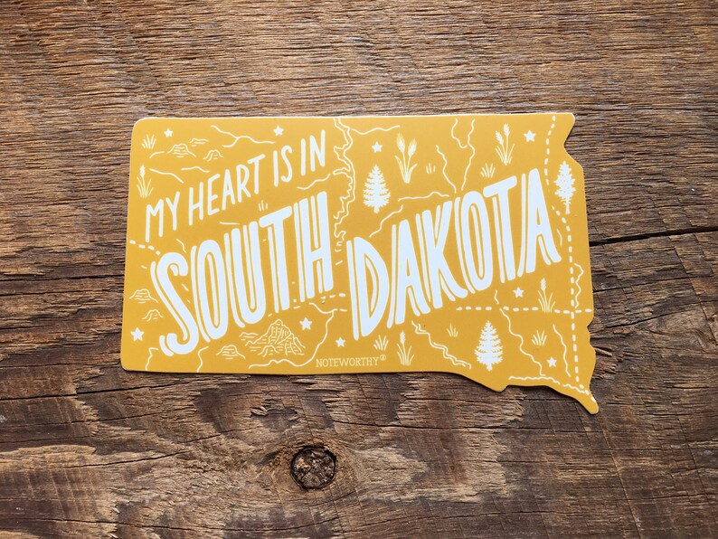 Pu&ograve; includere: Adesivo giallo a forma dello stato del Dakota del Sud con il testo "My heart is in South Dakota" e la parola "Noteworthy" in basso.