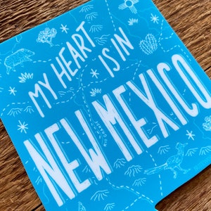 May include: Blue sticker with a white outline of a map of New Mexico. The text "My heart * is in NEW MEXICO" is written in white on the sticker. The sticker has a white star and a cactus on it.