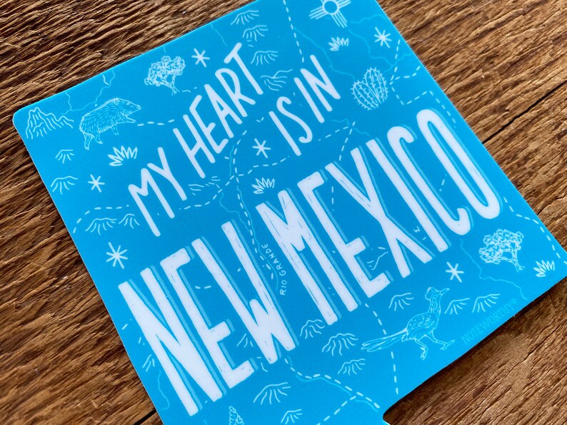 May include: Blue sticker with a white outline of a map of New Mexico. The text "My heart * is in NEW MEXICO" is written in white on the sticker. The sticker has a white star and a cactus on it.