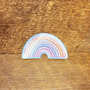 Pu&ograve; includere: Un cerotto di feltro a forma di arcobaleno con un contorno bianco. L'arcobaleno &egrave; composto da rosso, arancione, giallo, verde, blu, indaco e viola.