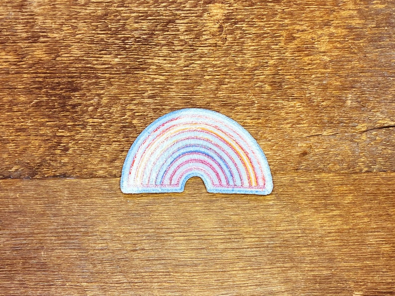 Pu&ograve; includere: Un cerotto di feltro a forma di arcobaleno con un contorno bianco. L'arcobaleno &egrave; composto da rosso, arancione, giallo, verde, blu, indaco e viola.