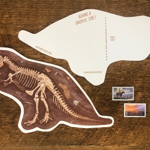 Peut inclure: Une carte postale illustrant un squelette de Tyrannosaurus Rex avec le texte "Having a Dinomite Time!" et "Tyrannosaurus Rex". La carte postale comporte un espace vide pour un message et une adresse, avec le texte "To:" et "Address goes to Postal Service here".