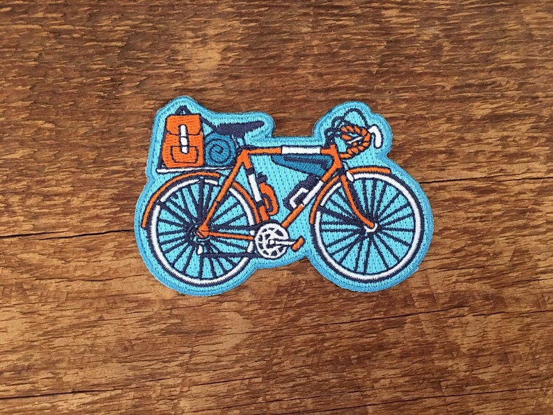 May include: An orange and blue embroidered patch of a bicycle with a backpack and sleeping bag attached to the back rack.