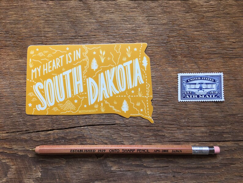Pu&ograve; includere: Un adesivo giallo con una mappa del Dakota del Sud e il testo "My heart is in South Dakota". L'adesivo &egrave; su una superficie di legno con un francobollo aereo blu e una matita marrone.