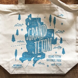 K&ouml;nnte beinhalten: Eine wei&szlig;e Canvas-Tragetasche mit einer blauen Grafik des Grand Teton National Park, Wyoming. Die Grafik enth&auml;lt eine Karte des Parks, Berge, B&auml;ume und die Worte "Grand Teton" in einer stilisierten Schrift. Der Text "Grand Teton National Park est. 1929" ist unter der Grafik gedruckt.