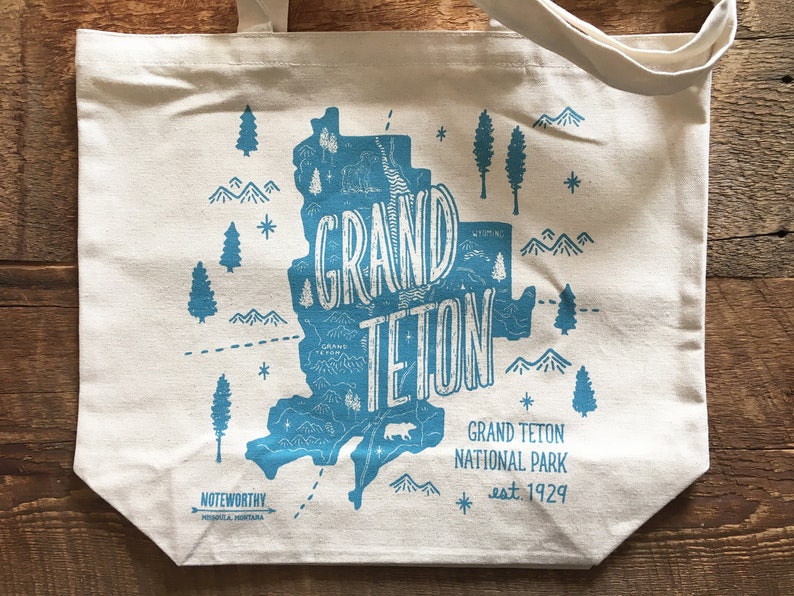 K&ouml;nnte beinhalten: Eine wei&szlig;e Canvas-Tragetasche mit einer blauen Grafik des Grand Teton National Park, Wyoming. Die Grafik enth&auml;lt eine Karte des Parks, Berge, B&auml;ume und die Worte "Grand Teton" in einer stilisierten Schrift. Der Text "Grand Teton National Park est. 1929" ist unter der Grafik gedruckt.