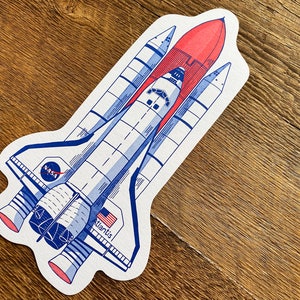 May include: A white and blue illustration of a space shuttle, the Atlantis, with the NASA logo on the side. The shuttle is depicted in flight with its engines firing and a red flame coming from the engine nozzle.