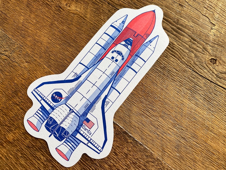 May include: A white and blue illustration of a space shuttle, the Atlantis, with the NASA logo on the side. The shuttle is depicted in flight with its engines firing and a red flame coming from the engine nozzle.