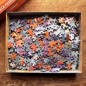 May include: A 1,000 piece jigsaw puzzle with a variety of colors, including orange, purple, and gray. The puzzle pieces are scattered in a box.