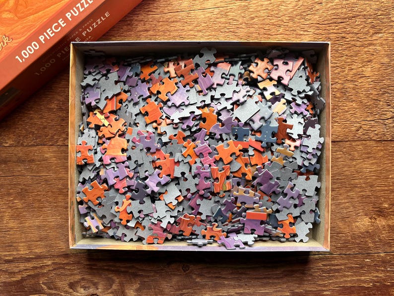 May include: A 1,000 piece jigsaw puzzle with a variety of colors, including orange, purple, and gray. The puzzle pieces are scattered in a box.