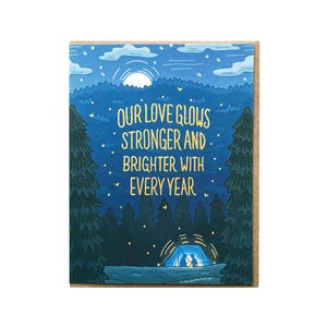 May include: An illustrated card in blue and green colours featuring a couple camping in a tent under a starry night sky. The text reads "Our love glows stronger and brighter with every year."