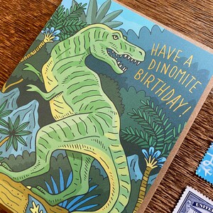 Op de afbeelding: Een groene dinosaurus met gele strepen staat in een jungle. De kaart luidt "Have a Dinomite Birthday!"