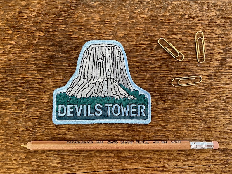 May include: A blue and green embroidered patch with a white outline depicting Devils Tower, a rock formation in Wyoming, USA. The text "Devils Tower" is embroidered in white on the patch.