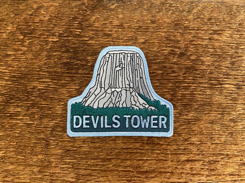 May include: A blue and green embroidered patch featuring a grey outline of Devils Tower National Monument with the text "Devils Tower" below.