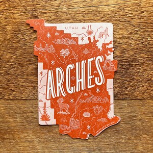 May include: A red and white illustrated map of Arches National Park in Utah. The map features a variety of landmarks, including the Delicate Arch, Balanced Rock, and the Colorado River. The word "Arches" is written in large, white letters across the map.