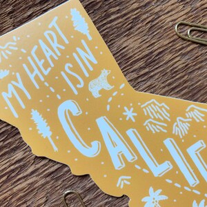 May include: A yellow outline of the state of California with the text "My heart is in California" in white lettering. The outline includes various illustrations of mountains, trees, a bear, and a star.