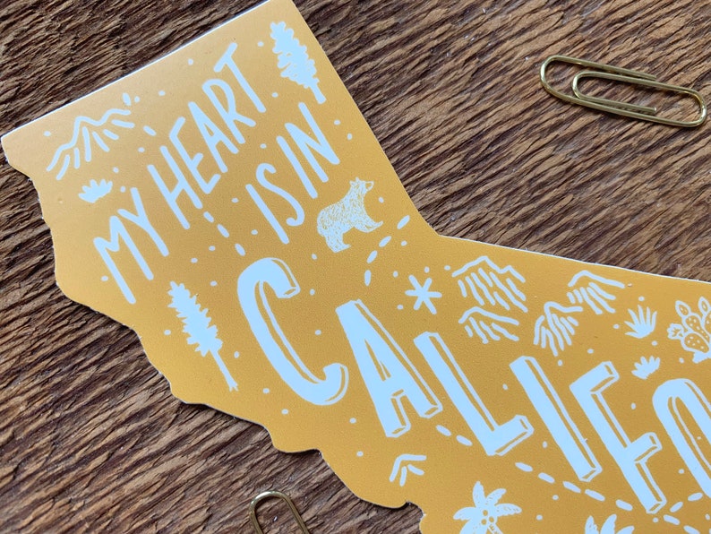 May include: A yellow outline of the state of California with the text "My heart is in California" in white lettering. The outline includes various illustrations of mountains, trees, a bear, and a star.