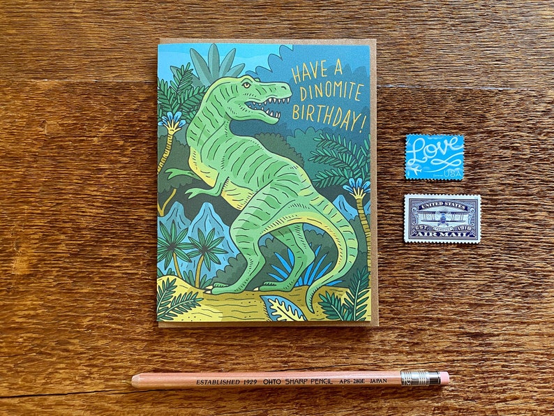 Op de afbeelding: Een groene dinosaurus met een tandenrij staat in een jungle-omgeving. De kaart luidt "Have a Dinomite Birthday!" en is versierd met groen, blauw en geel gebladerte.
