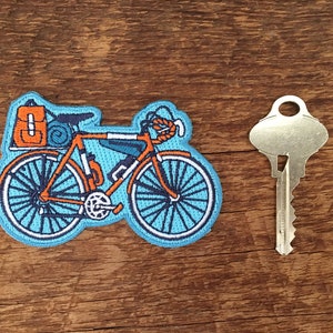 May include: A blue and orange embroidered patch of a bicycle with luggage attached to the handlebars and frame. The bicycle has a blue frame, orange handlebars, and white tires.