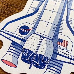 May include: A blue and white illustration of the Space Shuttle Atlantis with the NASA logo and the American flag. The text "Atlantis" is printed below the flag.