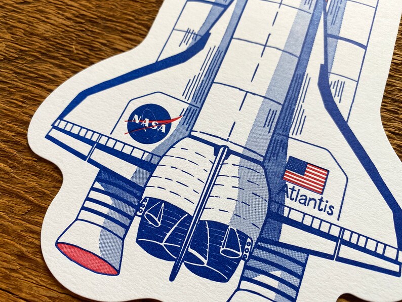 May include: A blue and white illustration of the Space Shuttle Atlantis with the NASA logo and the American flag. The text "Atlantis" is printed below the flag.