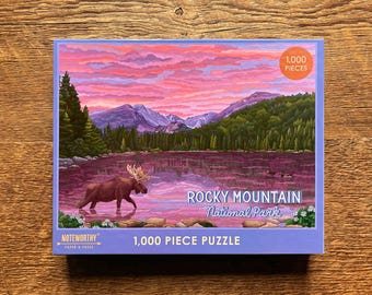 May include: A 1,000 piece jigsaw puzzle featuring a scenic view of Rocky Mountain National Park at sunset. The puzzle depicts a lake with a moose in the foreground and a mountain range in the background. The puzzle box is blue with white text and a picture of the completed puzzle.