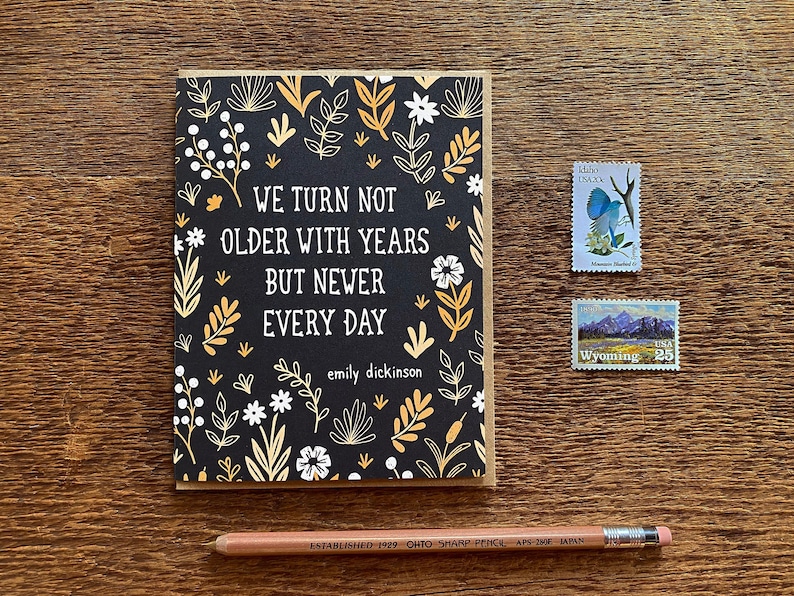May include: A black card with a floral pattern and the quote "We turn not older with years but newer every day" by Emily Dickinson.