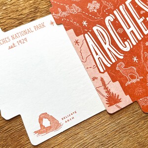 May include: A white postcard with a red outline shaped like the state of Utah. The postcard features a map of Arches National Park, Utah, with the text "Arches" in large, white letters. The postcard also includes the text "Arches National Park est. 1929" and "Delicate Arch".